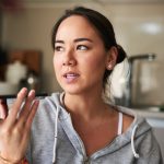 Eine Person in einem grauen Sweatshirt h&auml;lt ihr Smartphone in der Hand, ihr Blick schweift in die Ferne.