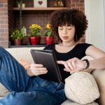 Eine Person liegt auf einem beigen Sofa. In den H&auml;nden h&auml;lt sie ein Tablet.