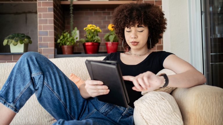 Eine Person liegt auf einem beigen Sofa. In den Händen hält sie ein Tablet.