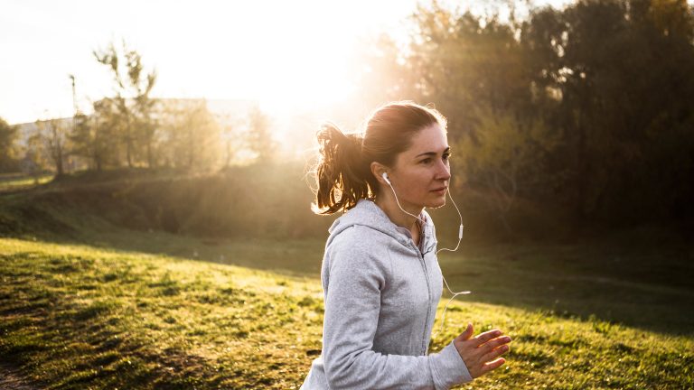 Eine Person joggt an einer Wiese entlang. Im Hintergrund ein paar Bäume und direkte Sonne.