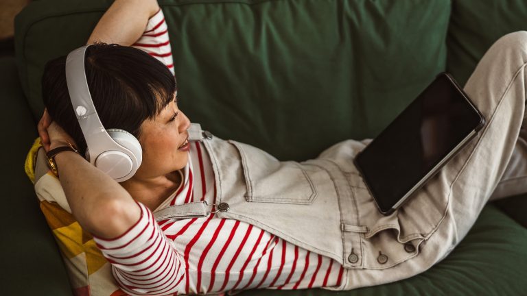 Eine Person liegt auf einer Couch und trägt Kopfhörer. Sie hat ein Tablet auf einem aufgestellten Bein abgestellt und die Arme hinter dem Kopf verschränkt.