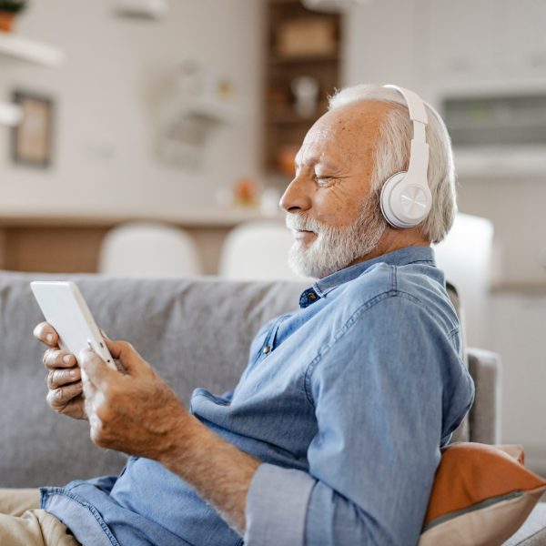 Eine &auml;ltere Person sitzt auf einem Sofa, schaut auf ihr Tablet und tr&auml;gt dabei Kopfh&ouml;rer.