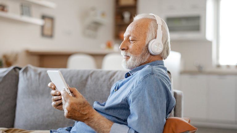 Eine ältere Person sitzt auf einem Sofa, schaut auf ihr Tablet und trägt dabei Kopfhörer.