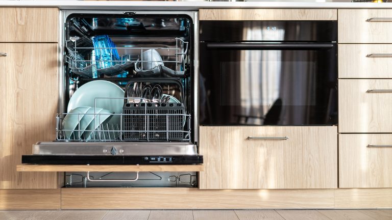 Ansicht einer holzfarbenen K&uuml;chenfront. Die Sp&uuml;lmaschine ist ge&ouml;ffnet. Es befinden sich t&uuml;rkise Teller und ein blaues Glas in der Maschine.