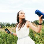 Eine Person steht auf einer Wiese. In einer Hand h&auml;lt sie einen Bluetooth-Lautsprecher, in der anderen Hand ein Smartphone.
