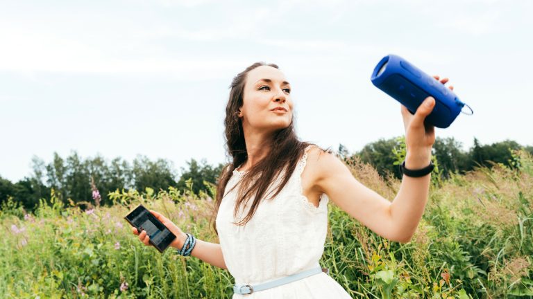 Eine Person steht auf einer Wiese. In einer Hand h&auml;lt sie einen Bluetooth-Lautsprecher, in der anderen Hand ein Smartphone.