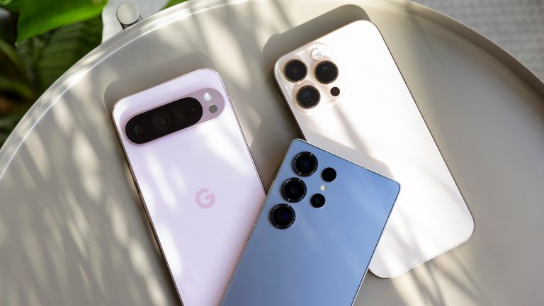 Ein Google Pixel Pro XL in Pink, ein Samsung Galaxy S25 Ultra in Blau und ein iPhone 16 Pro Max in Wüstensand liegen auf einem Tisch.