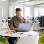 Eine Person sitzt in einem Gro&szlig;raumb&uuml;ro an einem runden Tisch, auf dem ein ge&ouml;ffnetes Notebook steht.