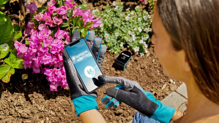 Eine Person sitzt vor einem Blumenbeet. Sie tr&auml;gt Gartenhandschuhe und h&auml;lt ein Smartphone in den H&auml;nden, auf dessen Display sich gerade die Gardena-App &ouml;ffnet.