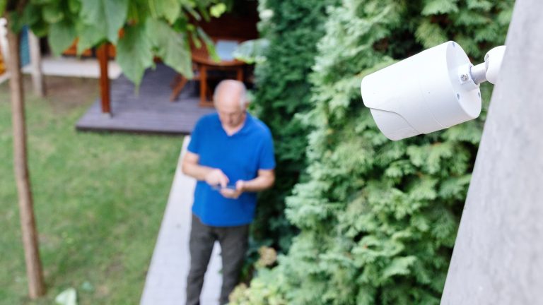 Im Fokus ist eine weiße Sicherheitskamera, die an einer Außenwand angebracht ist und auf den Garten gerichtet ist. Im Hintergrund ist eine Person zu sehen, die auf ein Smartphone tippt, das in ihren Händen liegt.