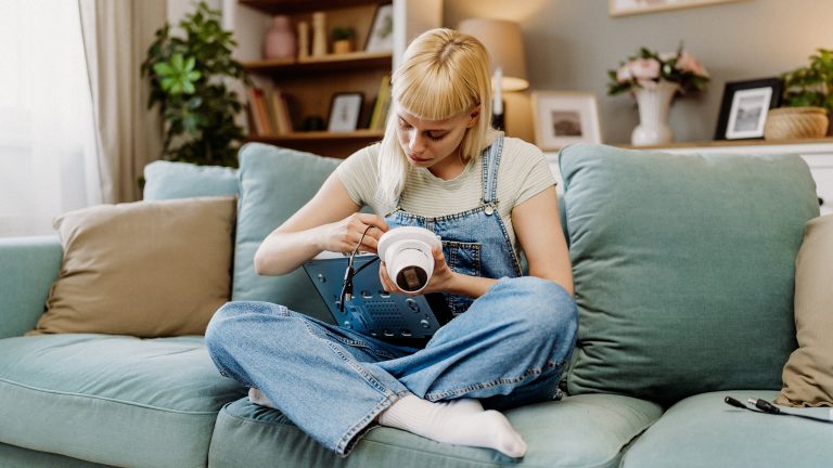 Eine Person sitzt im Schneidersitz auf einer Couch. Auf ihrem Schoß liegt eine Kamera mit einem Kabel. Sie nimmt diese gerade in Betrieb.
