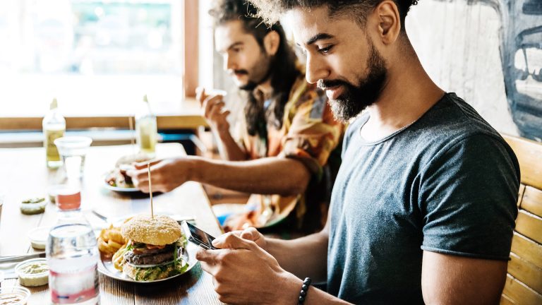 Zwei Personen sitzen am Tisch und Essen. Eine Person bedient dabei ihr Smartphone.