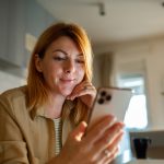 Eine Person h&auml;lt ein Smartphone in ihrer Hand, in welches sie hineinblickt. Im Hintergrund ist eine Kaffeetasse sowie ein Teil einer K&uuml;che zu erahnen.