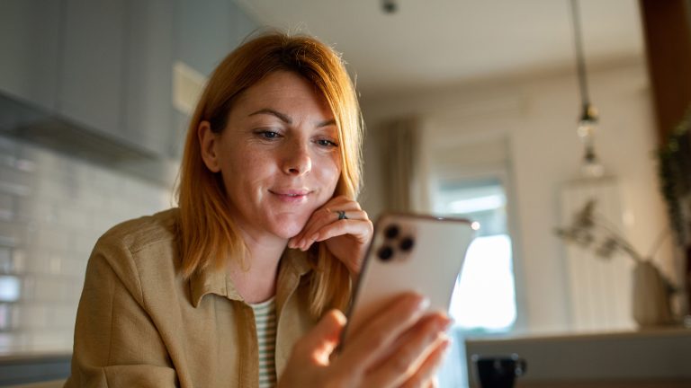 Eine Person hält ein Smartphone in ihrer Hand, in welches sie hineinblickt. Im Hintergrund ist eine Kaffeetasse sowie ein Teil einer Küche zu erahnen.