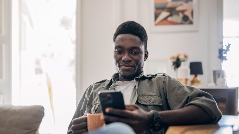 Eine Person sitzt auf einem Sofa. In der einen Hand hält sie eine Tasse, in der anderen ein Smartphone. Der Blick der Person ist auf das Smartphone gerichtet.