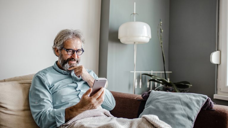 Eine Person sitzt bequem auf einer Couch. Auf ihrem Schoß liegt eine Decke, in der Hand hält sie ein Smartphone.