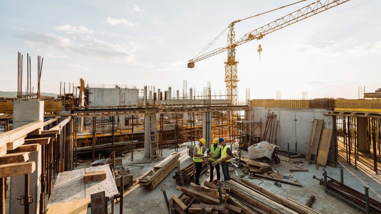 Drei Personen stehen zusammen auf einem Bauplatz und schauen in einen Plan. Sie sind umgeben von Stahlstangen. Im Hintergrund steht ein Kran.