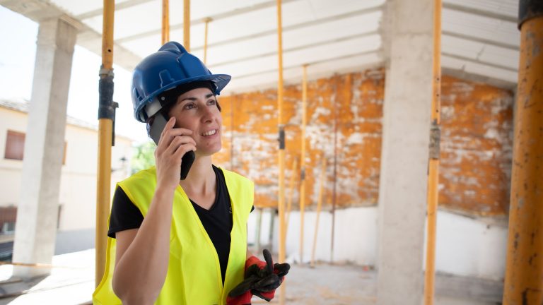Eine Person steht mit einer Sicherheitsweste und einem Schutzhelm bekleidet auf einer Baustelle und telefoniert mit ihrem Smartphone.