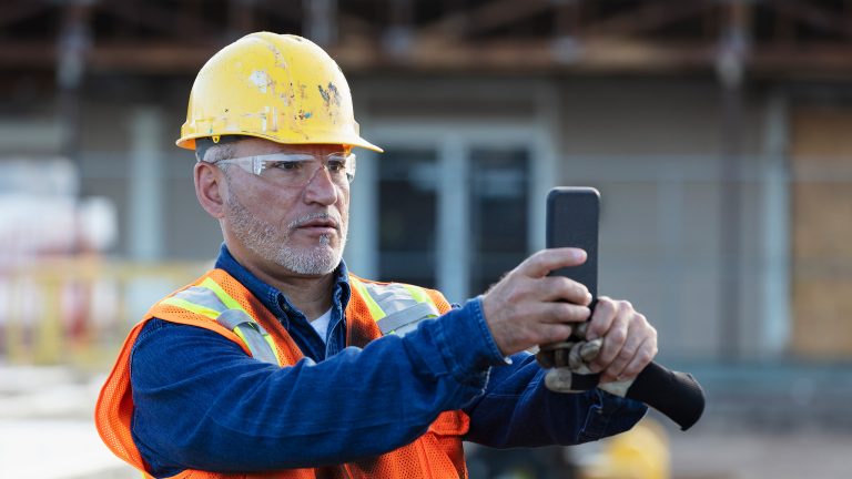 Eine Person mit einer Sicherheitsweste und einem Schutzhelm, hält ein Smartphone in den Händen.
