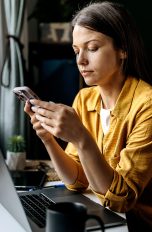 Eine Person sitzt an einem Schreibtisch, auf dem ein geöffneter Laptop steht. Sie schaut in ein Smartphone, das sie in beiden Händen hält.