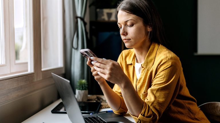 Eine Person sitzt an einem Schreibtisch, auf dem ein geöffneter Laptop steht. Sie schaut in ein Smartphone, das sie in beiden Händen hält.