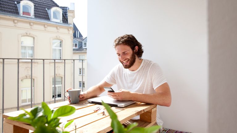 Eine Person sitzt vor einem zugeklappten Laptop und hat ein Smartphone in der Hand.