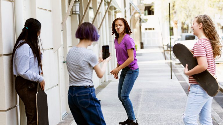 Eine Person steht auf einem Skateboard, eine weitere Person richtet ihr Smartphone auf sie und nimmt ein Video auf.