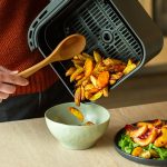Die Hand einer Person h&auml;lt einen Holzkochl&ouml;ffel, um Kartoffelwedges aus dem Frittierkorb eines Airfryers in eine Sch&uuml;ssel zu geben. Daneben steht ein Salat mit Pfirsichvierteln.