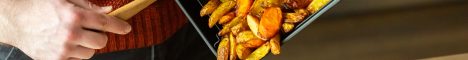 Die Hand einer Person hält einen Holzkochlöffel, um Kartoffelwedges aus dem Frittierkorb eines Airfryers in eine Schüssel zu geben. Daneben steht ein Salat mit Pfirsichvierteln.