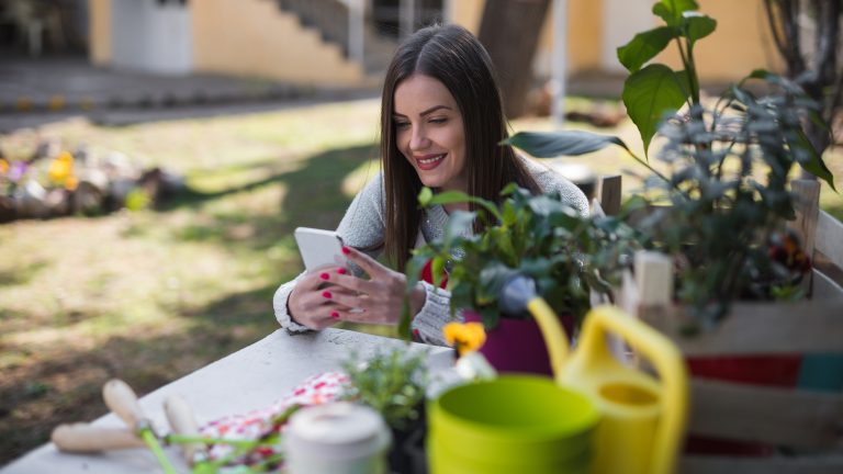 Eine Person sitzt im Garten an einem Tisch, auf dem verschiedene Werkzeuge zur Gartenarbeit liegen. In den H&auml;nden h&auml;lt sie ein Smartphone.