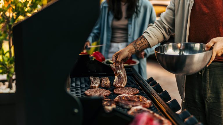 Seitenansicht eines Gasgrills auf dem bereits Steaks und Pattys liegen. Eine Person, die eine Sch&uuml;ssel in der linken Hand h&auml;lt, ist gerade dabei mit der rechten Hand ein weiteres Steak dazu zu legen.