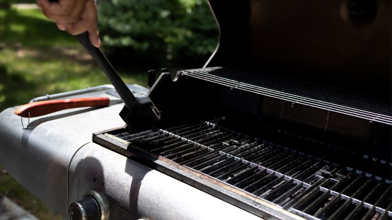 Es ist die Hand einer Person zu sehen, die mit einer Eisenb&uuml;rste grobe Reste vom Grillrost entfernt.