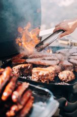 So kannst du deinen Gasgrill reinigen Auf dem Grillrost liegen zahlreiche Fleischstücke. Flammen bersten durch den Rost, darüber eine Hand mit einer Zange, um das Fleisch zu wenden.