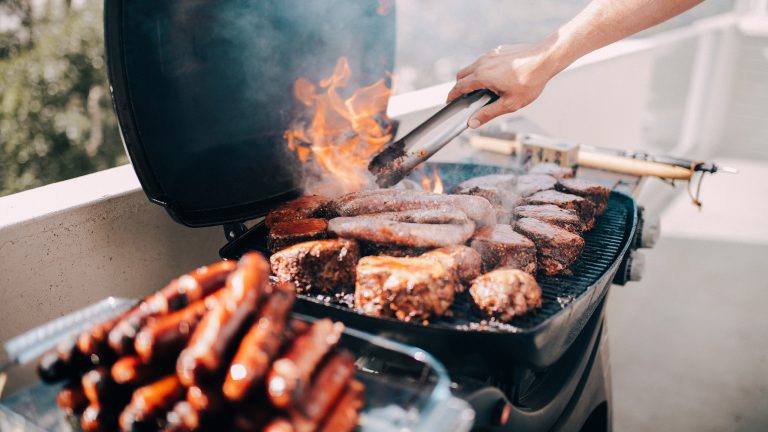 Auf dem Grillrost liegen zahlreiche Fleischst&uuml;cke. Flammen bersten durch den Rost, dar&uuml;ber eine Hand mit einer Zange, um das Fleisch zu wenden.