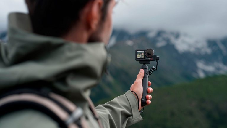 Eine Person trägt die Osmo Action 5 Pro Adventure Combo Action Cam auf einem Selfie-Stick. Im Hintergrund sind schneebedeckte Berge zu sehen.