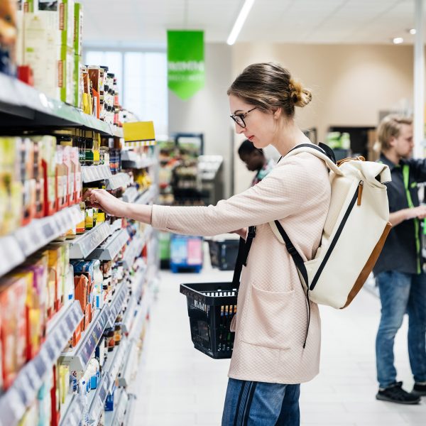 Eine Person steht vor einem Supermarktregal, aus dem sie gerade etwas herausnehmen m&ouml;chte. In der anderen Hand h&auml;lt sie einen Einkaufskorb.