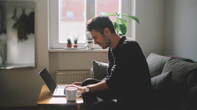 Eine Person sitzt auf einem Sofa in einem Wohnzimmer und tippt auf einem Laptop, der vor ihr auf einem kleinen Tisch steht.