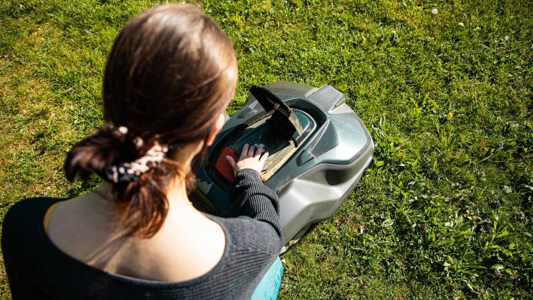 Mähroboter ohne Begrenzungskabel, Darauf kommt es an Eine Person kniet vor einem Mähroboter. Eine Klappe des Geräts ist geöffnet damit die Person die Sensoren auf Funktionstüchtigkeit prüfen kann.