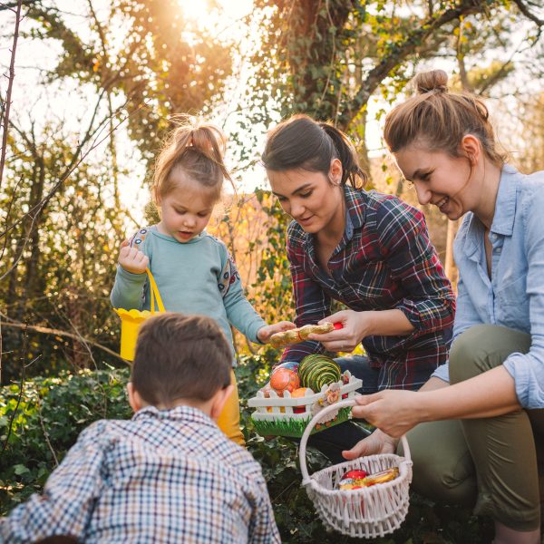 Zwei Personen halten jeweils ein kleines Osternest in den H&auml;nden, w&auml;hrend zwei Kinder im Garten nach Ostereiern suchen.