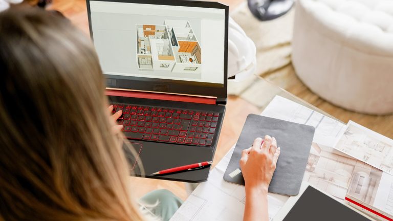 Eine Person sitzt an einem Glas-Schreibtisch. Auf dem Laptop vor ihr ist ein 3D-Grundriss eines Raumes zu sehen. Daneben liegen jede Menge Zettel mit Zeichnungen.