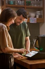 Thermomix TM7 Zwei Personen stehen in der Küche an einem Tisch, auf dem der neue TM7 steht. Eine der beiden Personen bedient diesen per Touchscreen.