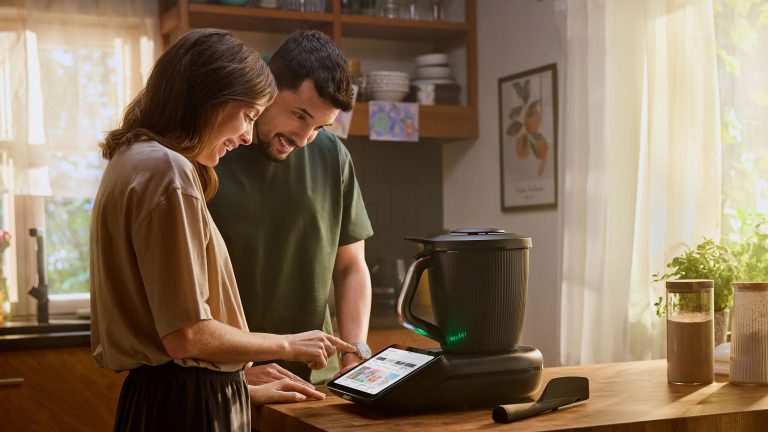 Zwei Personen stehen in der Küche an einem Tisch, auf dem der neue TM7 steht. Eine der beiden Personen bedient diesen per Touchscreen.
