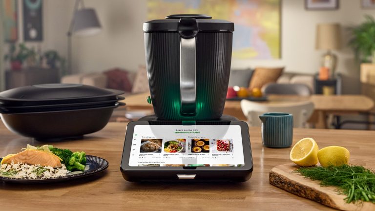 Der TM7 steht auf einer hölzernen Arbeitsplatte. Daneben steht ein Teller mit einer fertigen Reis-Speise sowie ein Holz-Schneidbrett mit Dill und einer aufgeschnittenen Zitrone.