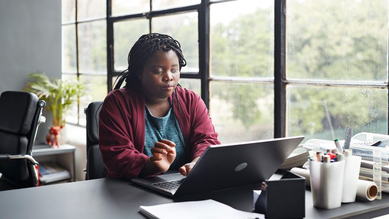 Eine Person mit einer Sehbeeinträchtigung sitzt in einem Büro an einem Schreibtisch vor einem geöffneten Notebook und tippt etwas auf der Tastatur.