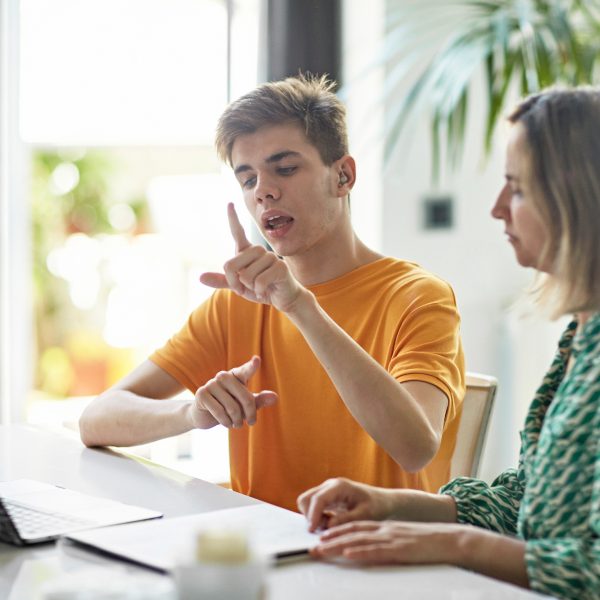 Zwei Personen sitzen an einem Tisch und schauen gemeinsam in einen Laptop. Eine der beiden Personen kommuniziert w&auml;hrenddessen mit ihren H&auml;nden in Geb&auml;rdensprache.
