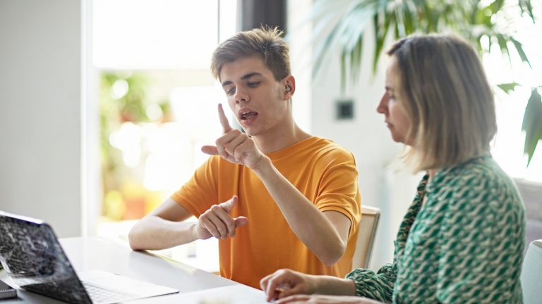 Zwei Personen sitzen an einem Tisch und schauen gemeinsam in einen Laptop. Eine der beiden Personen kommuniziert währenddessen mit ihren Händen in Gebärdensprache.