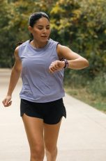 Eine Person in Trainingsklamotten l&auml;uft auf einem Weg durch einen Park. Sie blickt auf die Smartwatch an ihrem linken Handgelenk.