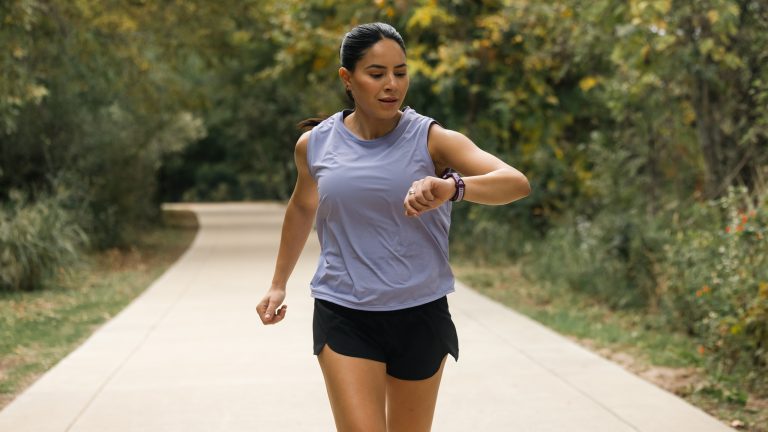 Eine Person in Trainingsklamotten läuft auf einem Weg durch einen Park. Sie blickt auf die Smartwatch an ihrem linken Handgelenk.