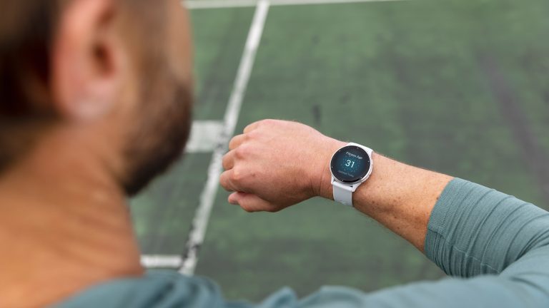 Blick über die Schulter einer Person: Sie steht am Rand eines Sportplatzes und blickt auf die Smartwatch an ihrem rechten Handgelenk.
