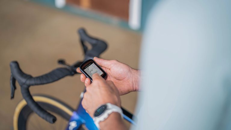 Eine Person hält einen Fahrradcomputer in den Händen. Im Hintergrund ist ein Gravelbike zu sehen.